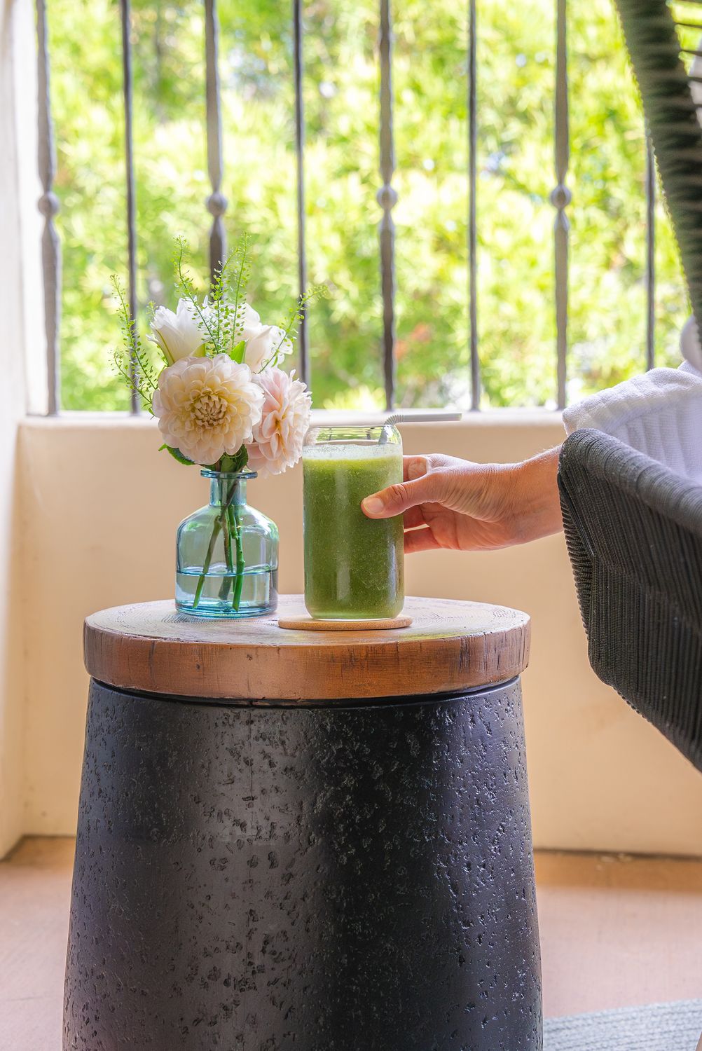 Hand holding green smoothie near flowers.